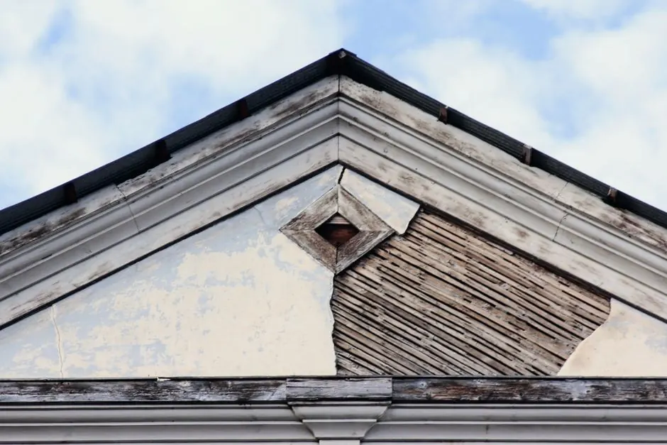 Close-up of a weathered building facade with peeling paint and exposed wooden planks against a blue sky.